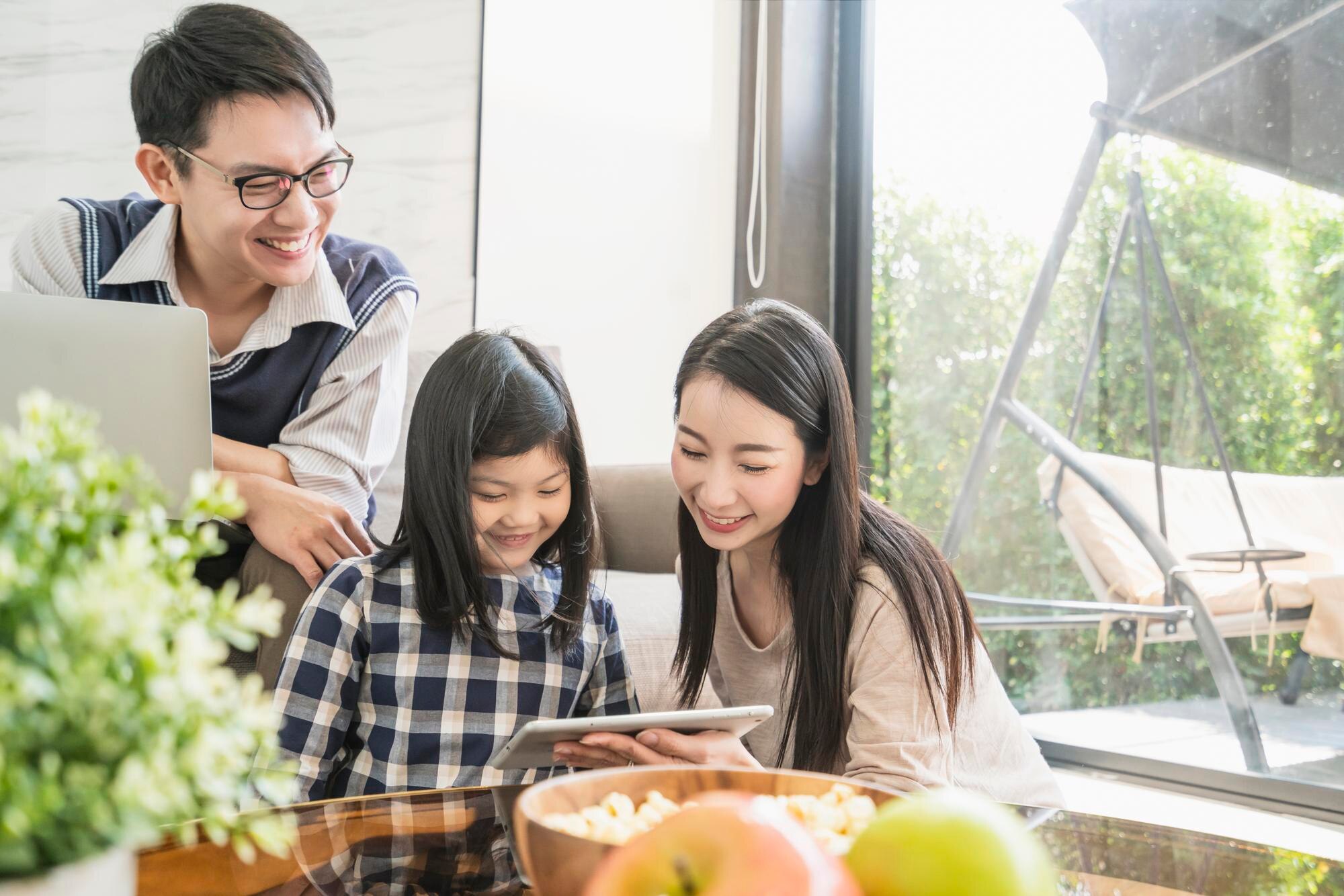 asian-daughter-is-doing-homework-notebook-his-mother-father-are-sitting-watch-sofa-use-digital-laptop-together-home-background_609648-1897